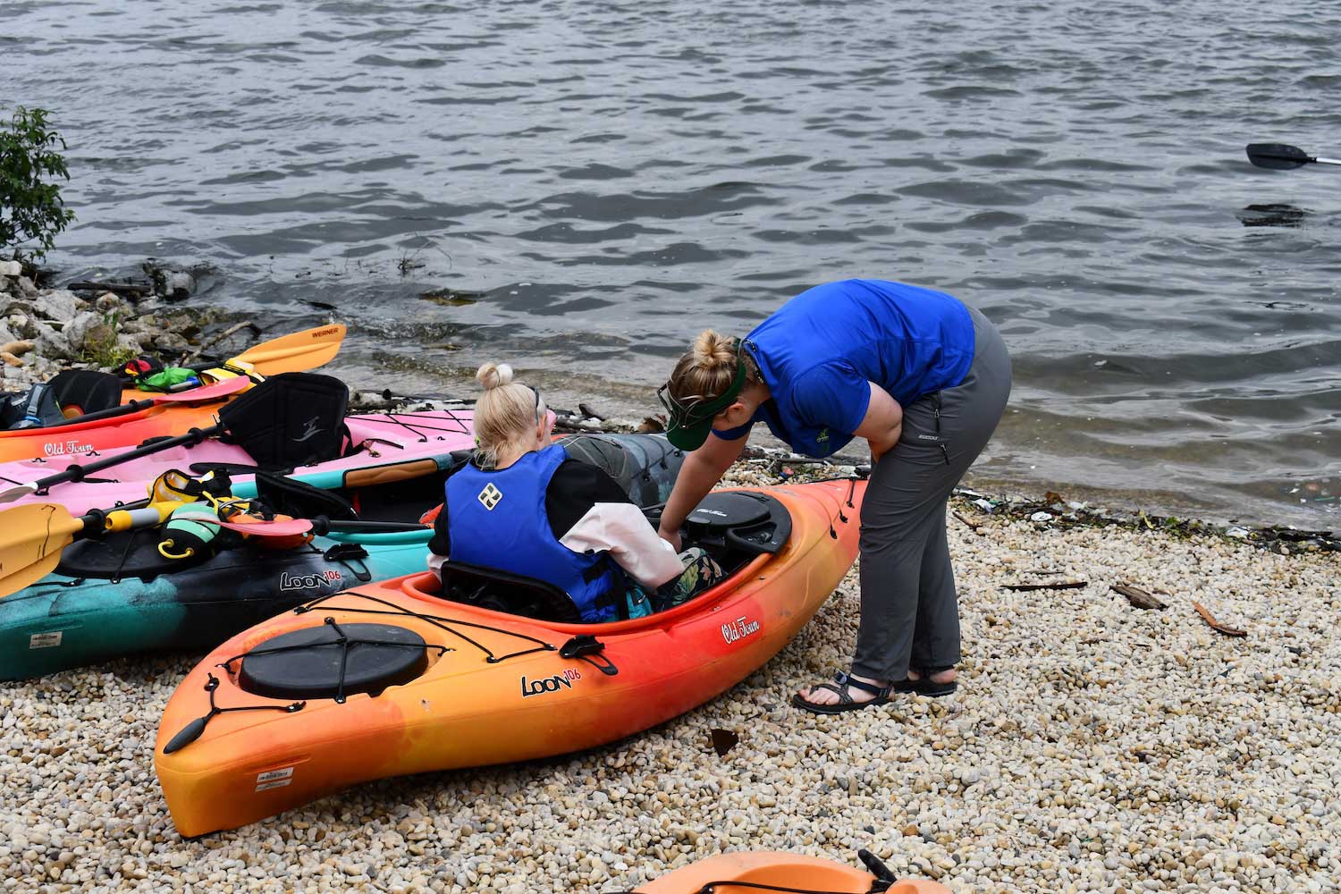 A person helping someone with a kayak with a group of kayaks situated at the water's edge.