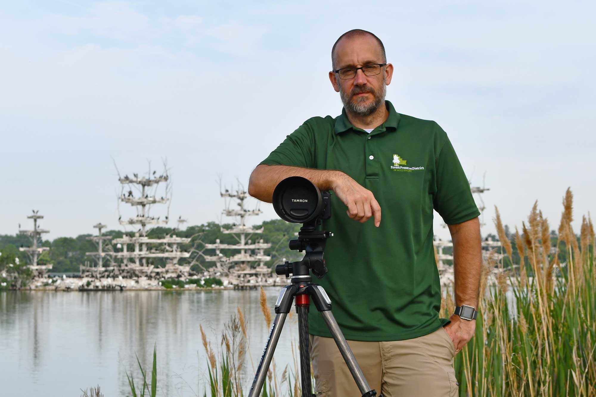 A man stands with a camera with a lake in the background.