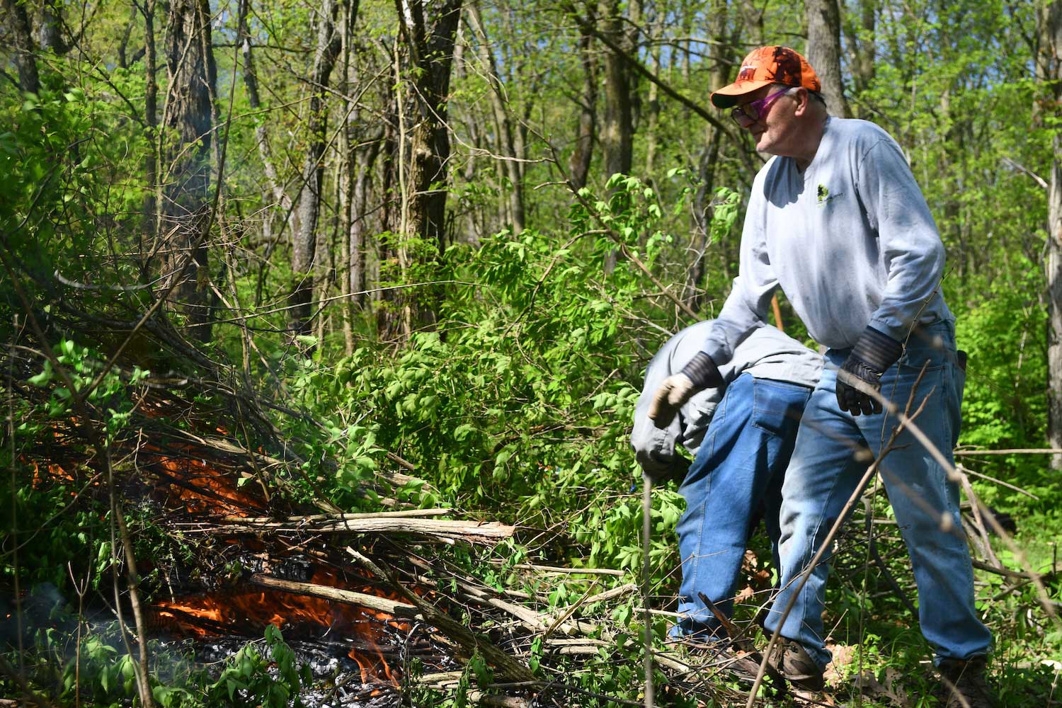 A person standing next to a burning brush pile while another person works crouched over in the background.