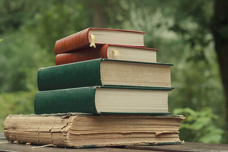A stack of books on a wooden table with trees in the background.