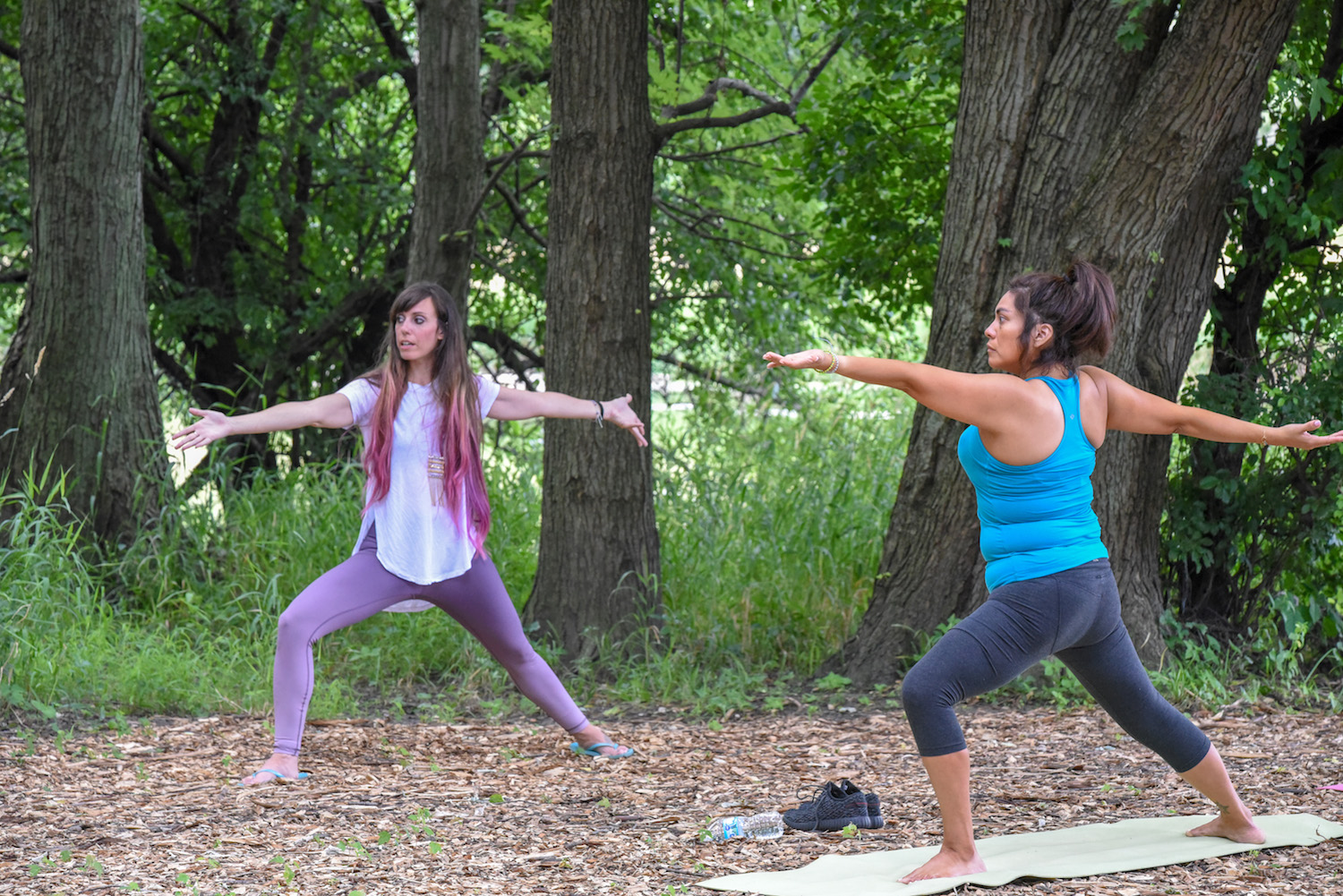 Two people doing yoga outdoors surrounded by big trees.