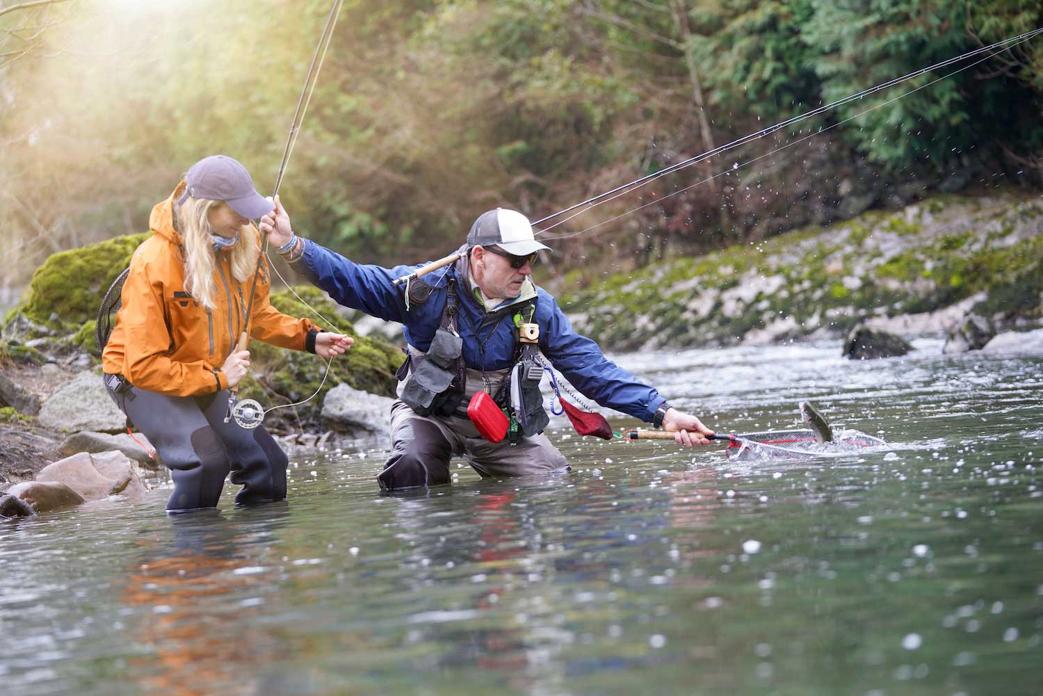 Two people wearing waders in the water while fly fishing, with one scooping up a fish in a net.