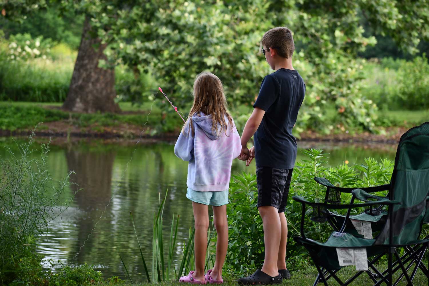 Two children holding fishing poles standing along the shore of a pond. 
