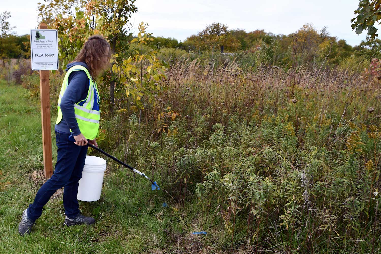 A person holding a bucket and grabbers will picking up litter along a trail.