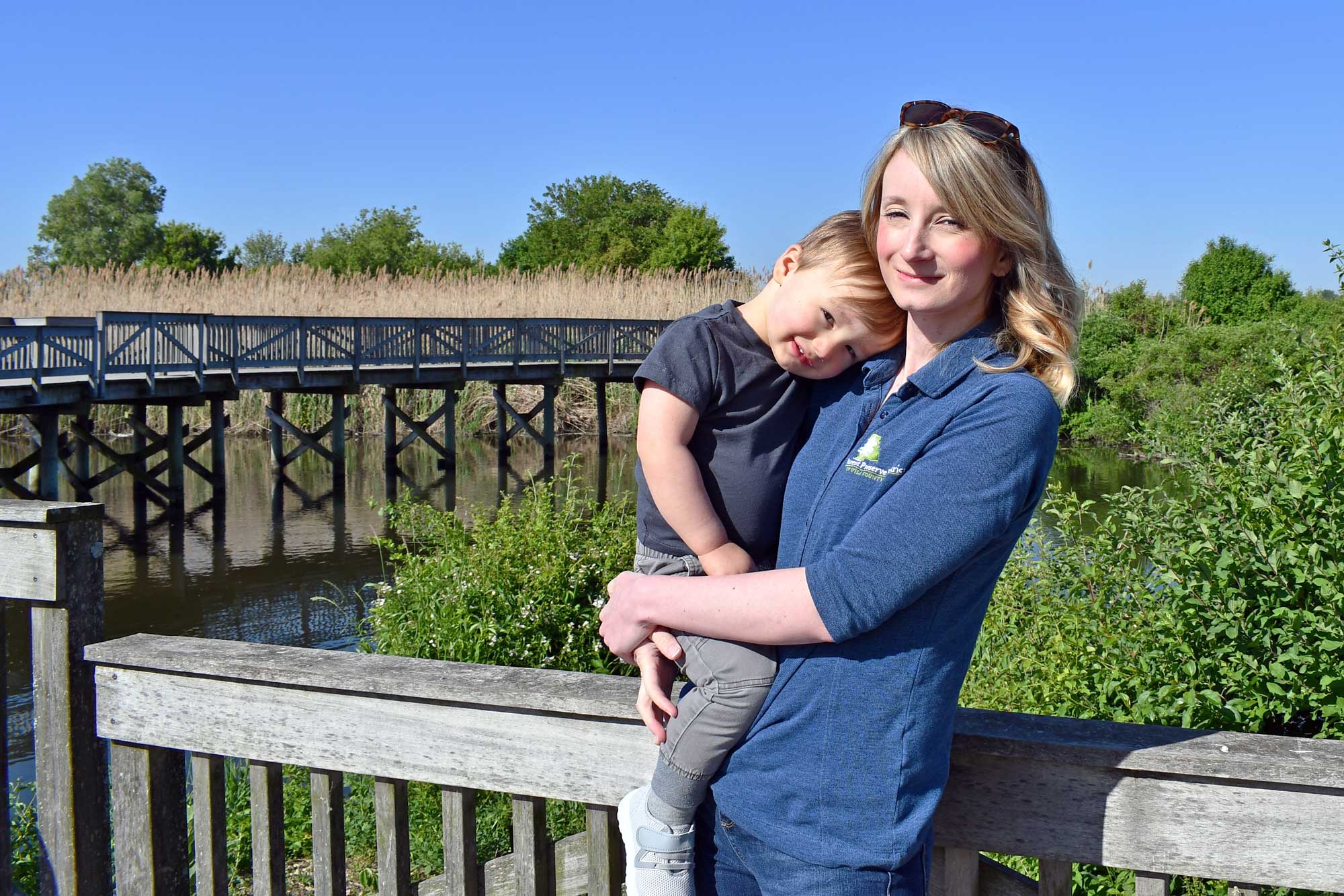 A woman holds her young child while on a long wooden bridge.