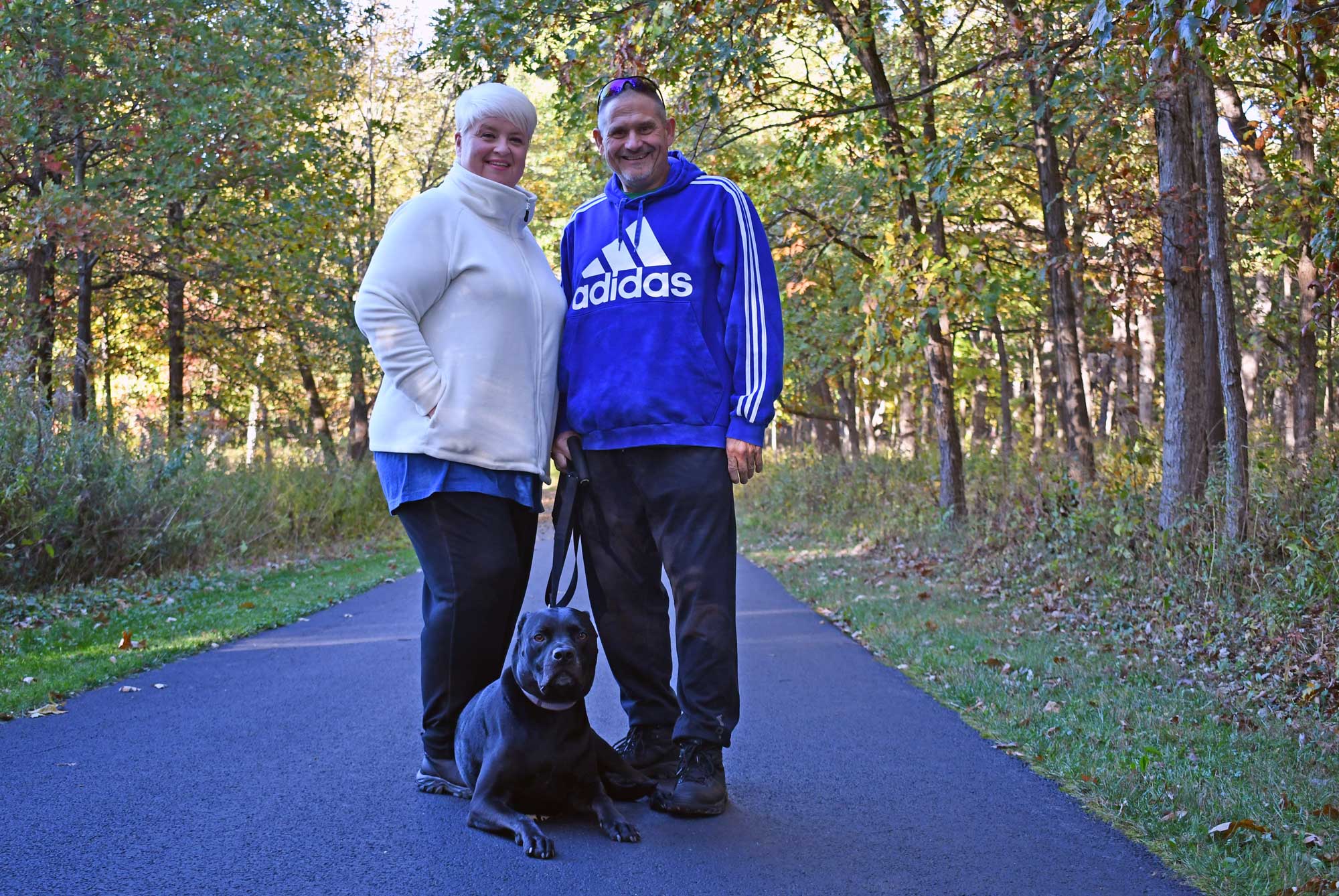 A man, woman, and their dog pose for a photo along a tree-lined trail.