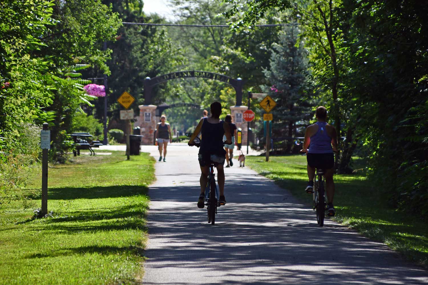 People walking and riding bikes along the Old Plank Road Trail on a sunny summer day.