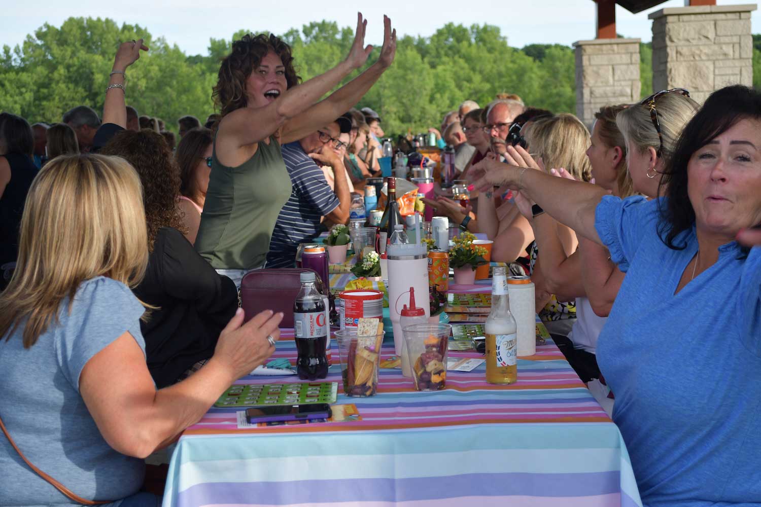 People playing bingo while sitting at picnic tables under a picnic shelter. One person is standing up to announce they have won. 